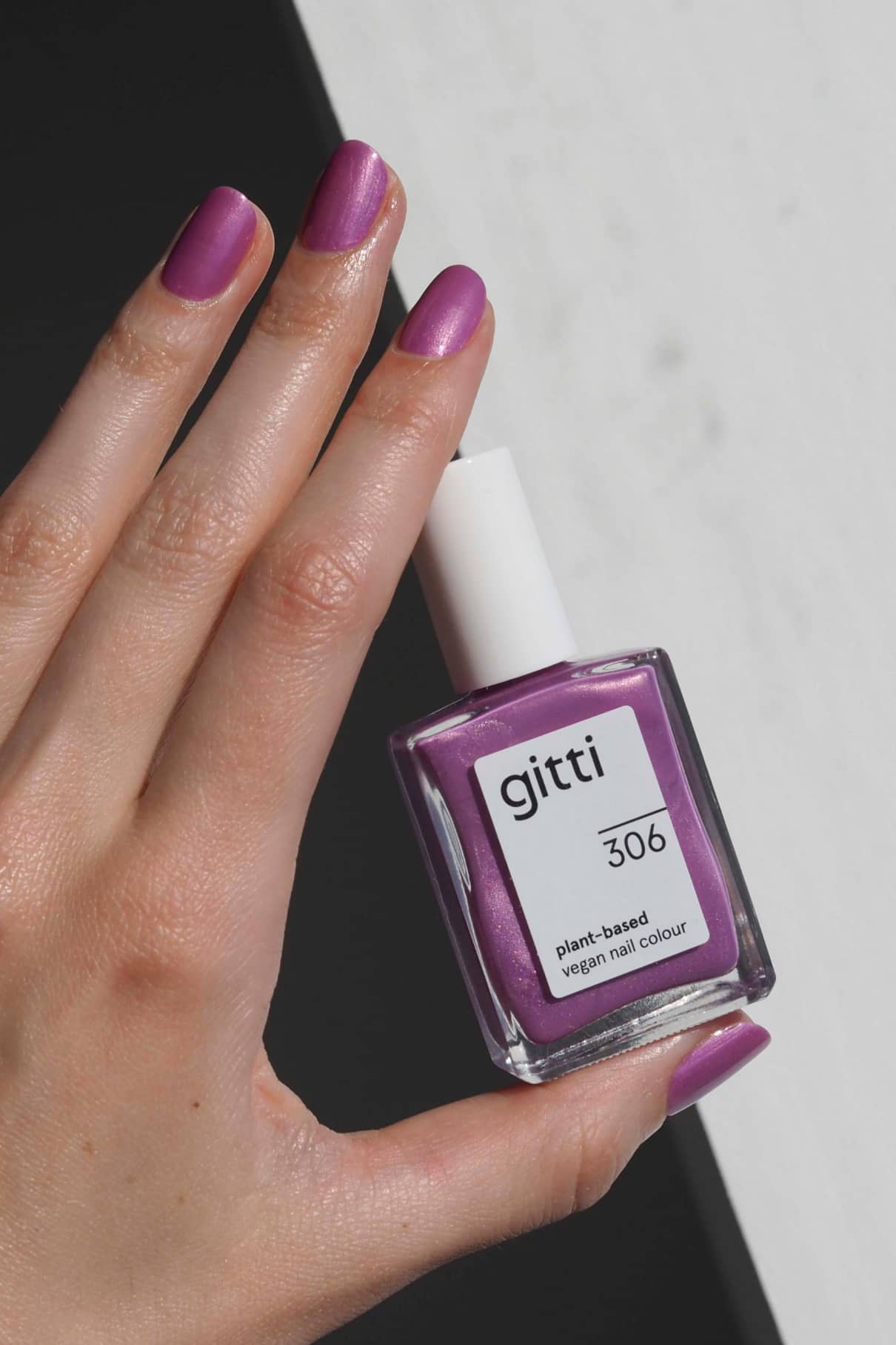 Eine Hand mit lilafarbenen Nägeln mit einem violetten Unterton hält eine Flasche gitti no. 306 pflanzlicher veganer Nagellack vor einem schwarz-weißen Hintergrund.