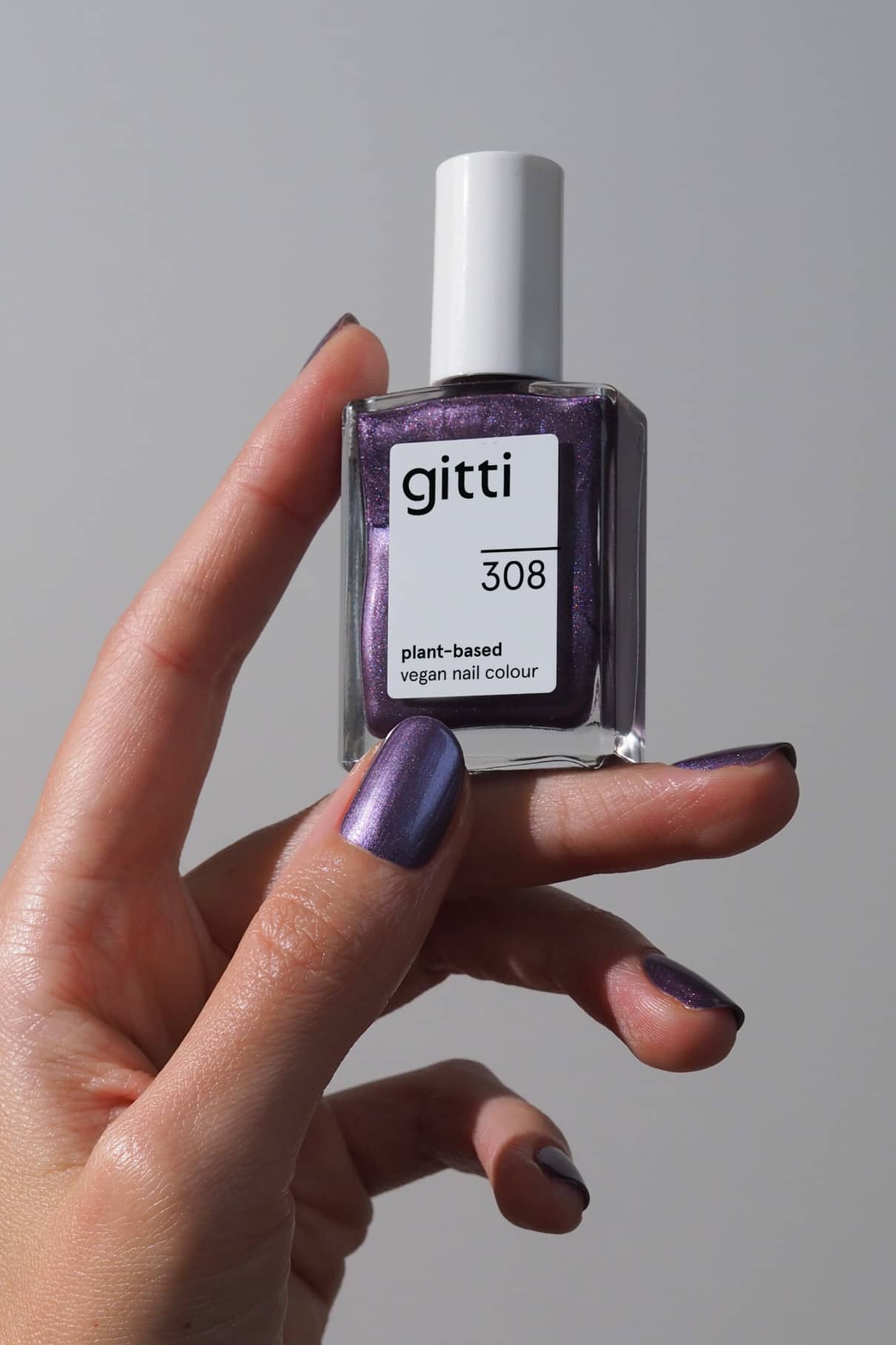 Eine Hand mit metallisch schimmernden lila Nägeln hält eine Flasche gitti no. 308 pflanzlichen, veganen Nagellack im Farbton Violettton vor einem schlichten, hellen Hintergrund.