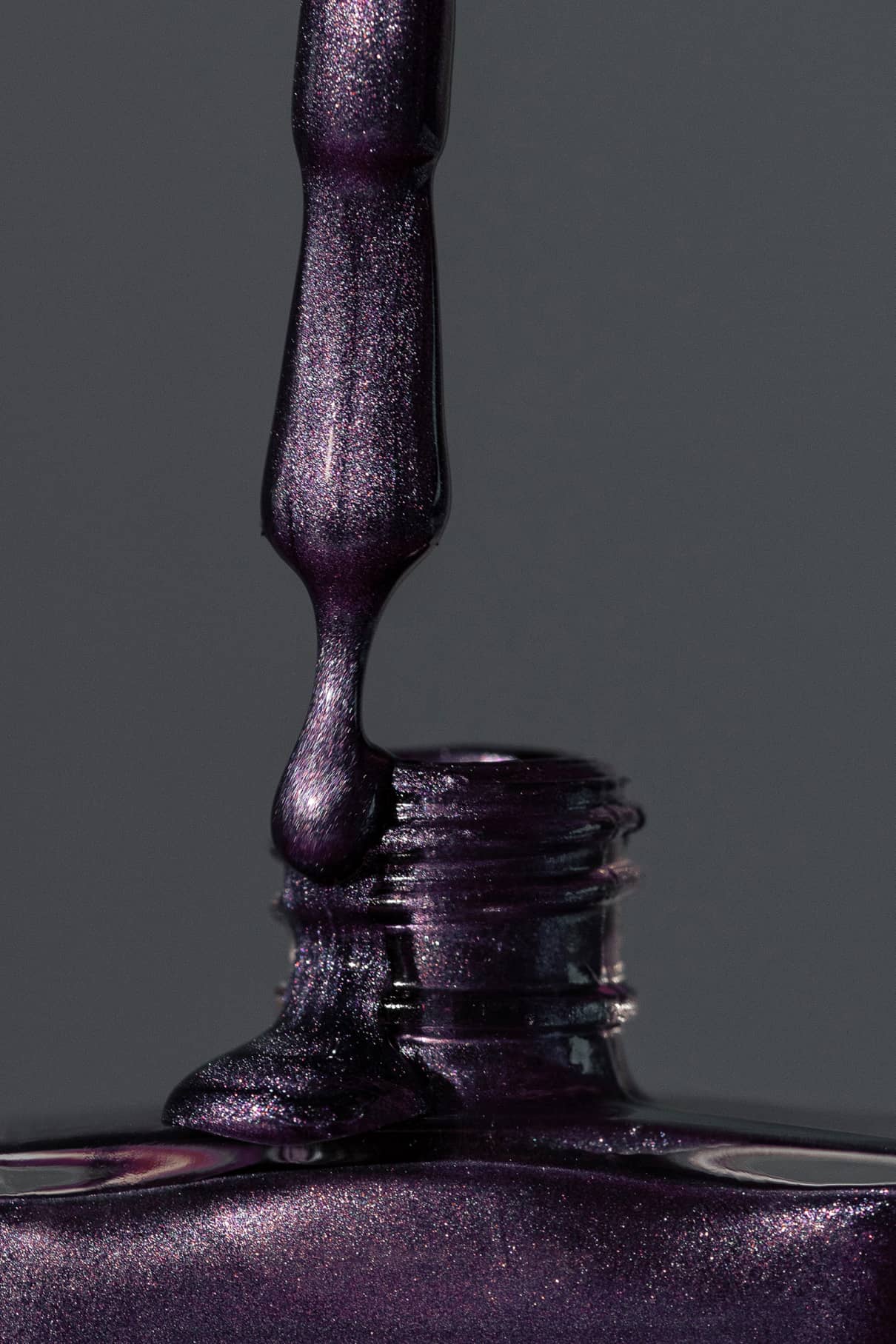 Eine Nahaufnahme eines Nagellackpinsels, der gitti no. 308 dunkelviolett schimmernder Metallic-Lack in eine offene Flasche vor einem grauen Hintergrund.