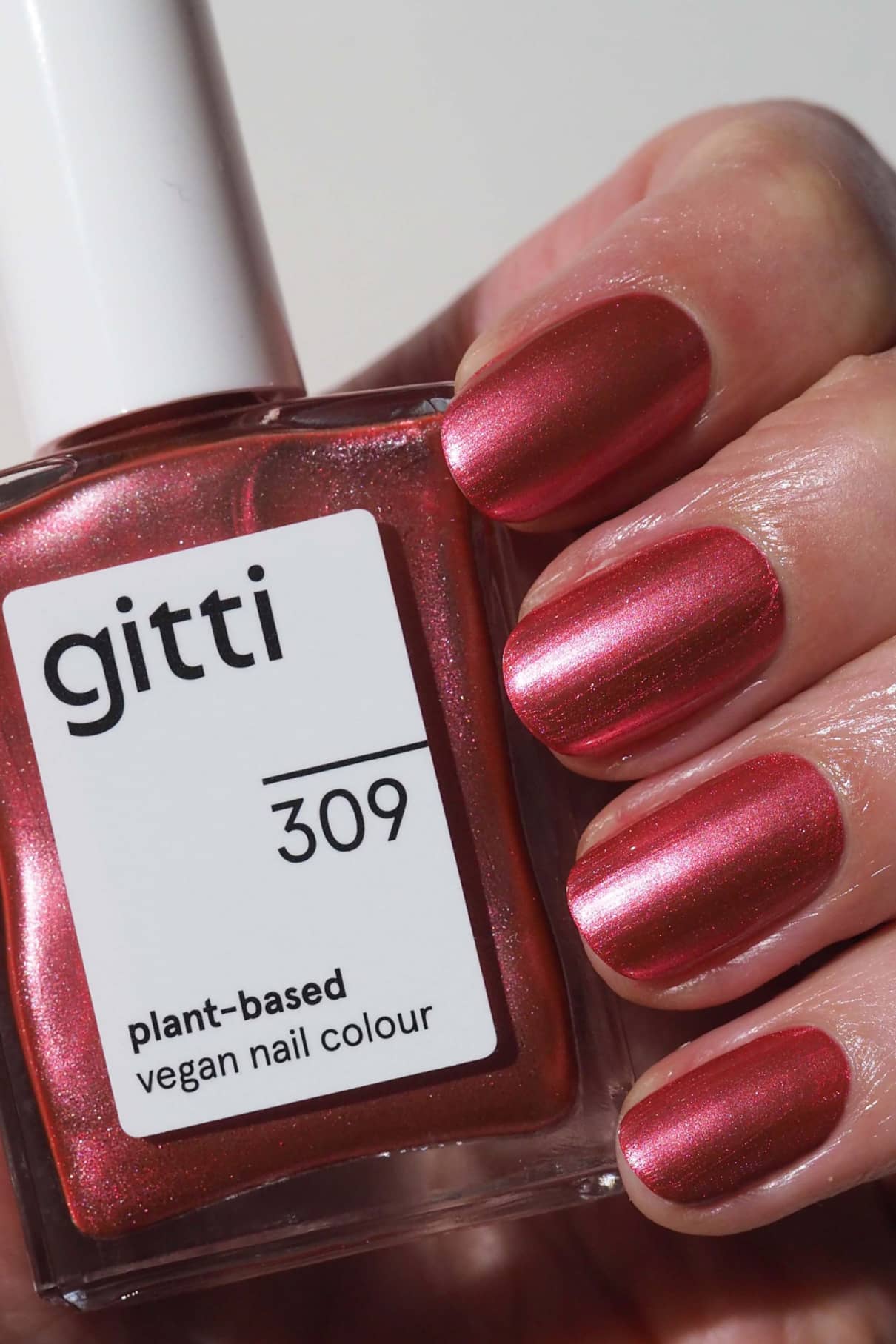 Eine Hand mit kurzen, in Pink Metallic lackierten Nägeln hält ein Fläschchen mit gitti no. 309 pflanzlicher veganer Nagellack in rötlichem Pink.
