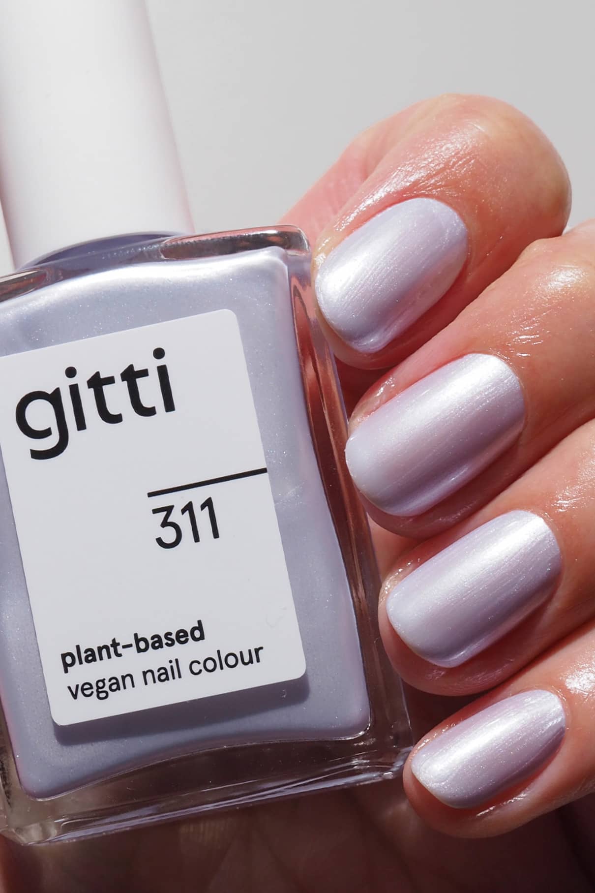 Eine Hand mit kurzen, in einem perlmuttfarbenen Lila-Ton lackierten Nägeln hält eine Flasche gitti no. 311, einer pflanzlichen veganen Nagelfarbe in einem kühlen blauen Metallic-Finish, das zum Nagelton passt.