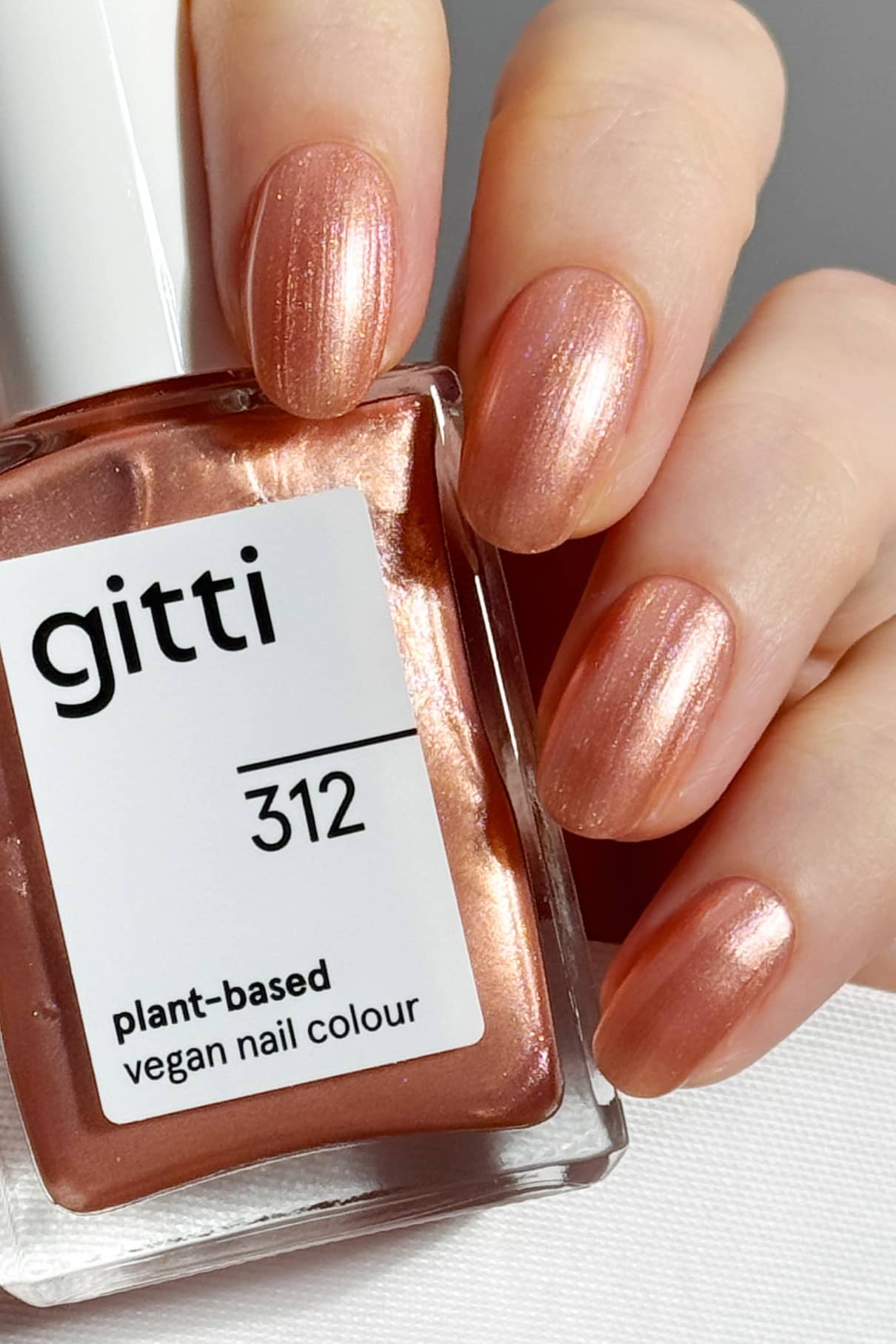 Eine Hand mit kurzen, in schimmerndem Roségold lackierten Nägeln hält ein Fläschchen mit der Aufschrift Quick Dry Routine - pflanzliche, vegane Nagelfarbe, die mit einem High Gloss Top Coat für zusätzlichen Glanz versehen ist.