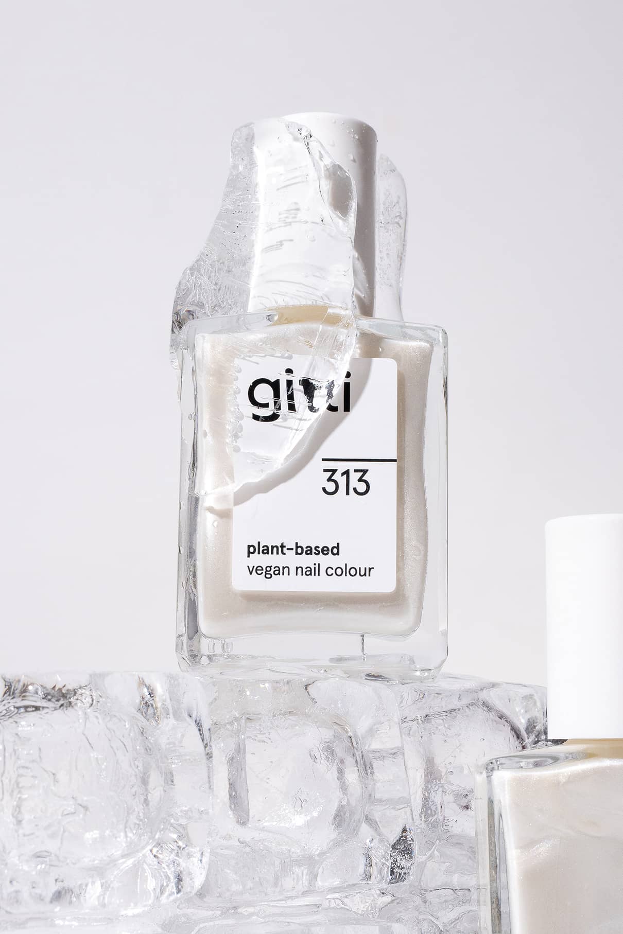Ein Fläschchen von gitti no. 313, eine pflanzliche, vegane Nagelfarbe in Metallic-Weiß, ruht auf Eis mit einem schmelzenden Fragment - perfekt für den Trendlook dieser Saison.