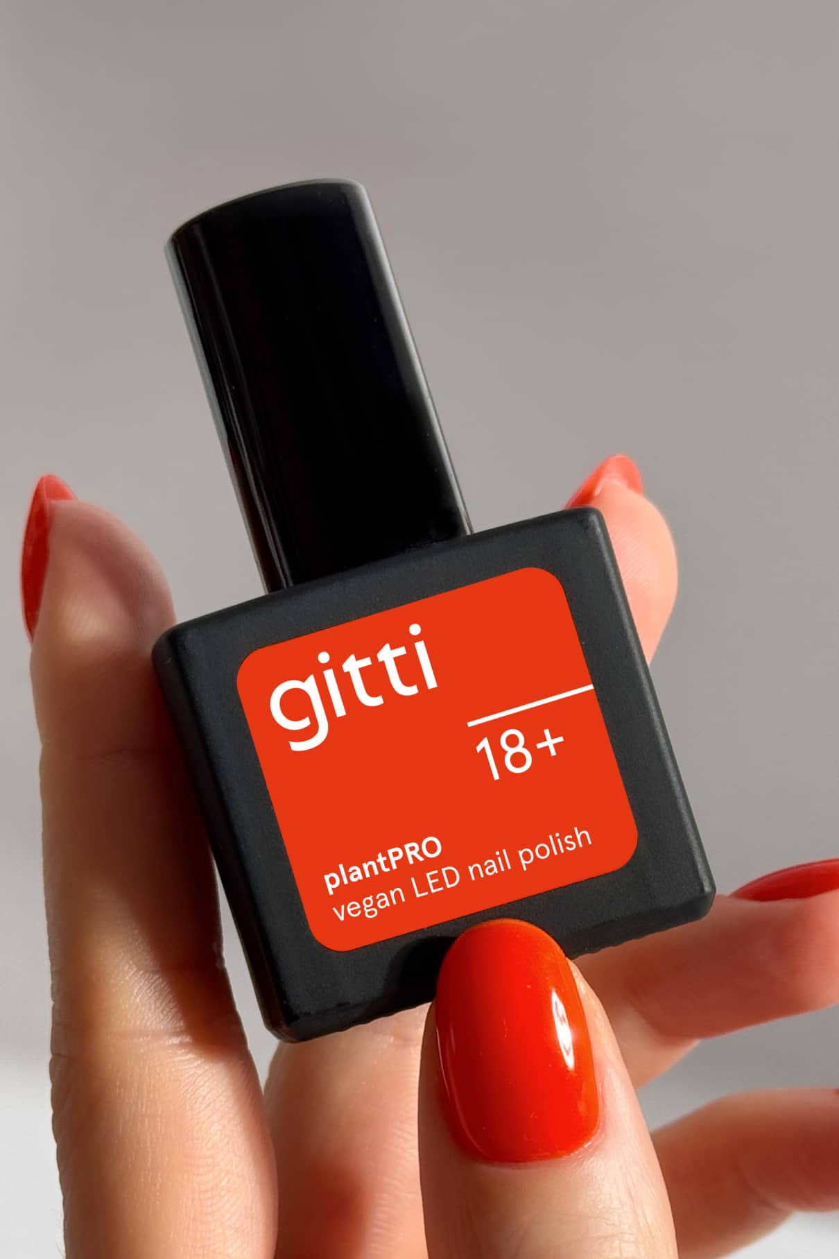 Eine Hand mit rot lackierten Nägeln in leuchtendem Orange-Rot hält eine schwarze Flasche mit der Aufschrift gitti no. 18+ plantPRO vegan LED Nagellack.