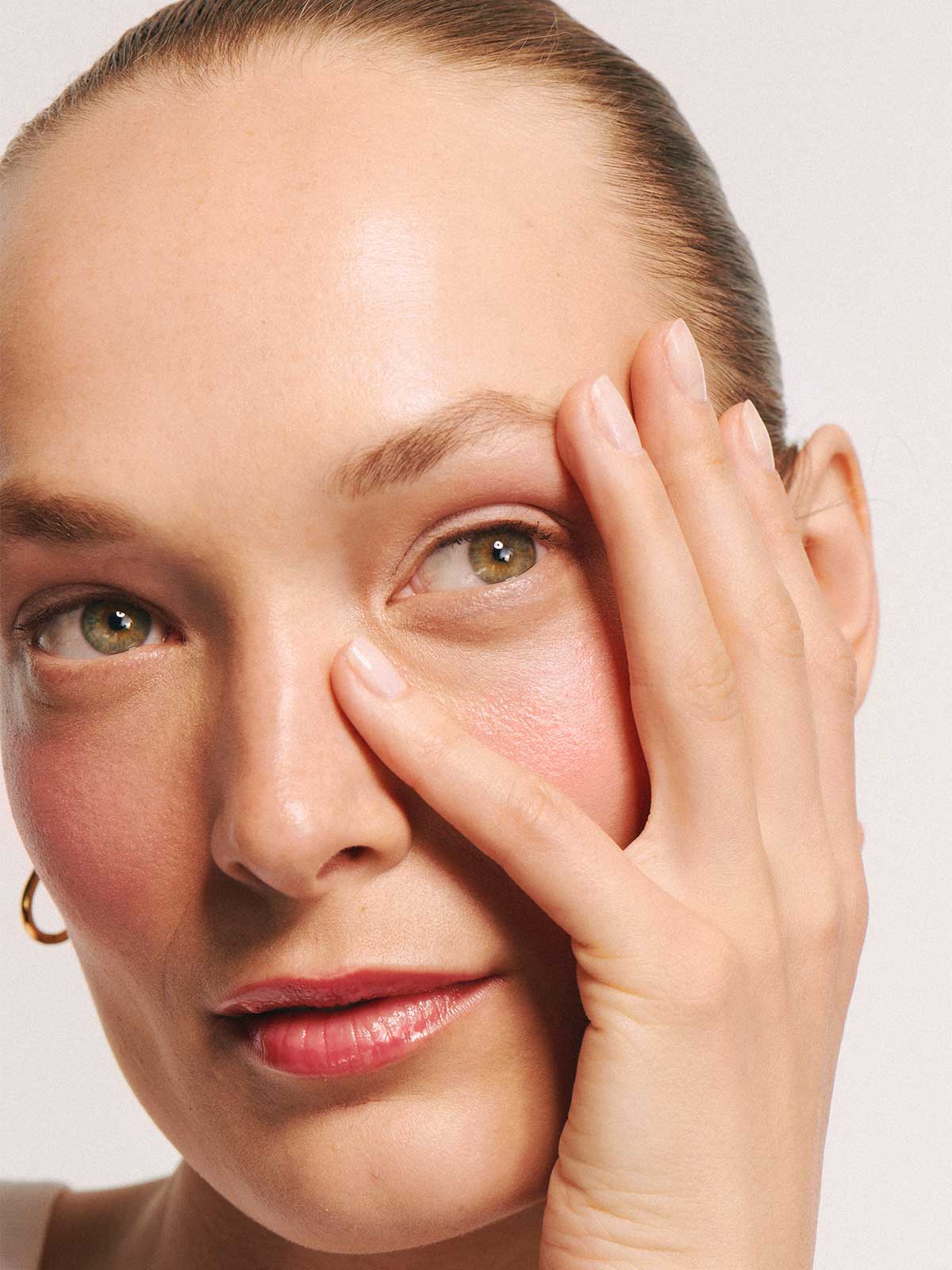 Eine Person mit hellen Augen und heller Haut blickt nach vorne und berührt ihr Gesicht mit der Hand vor einem neutralen Hintergrund, wodurch der sanfte Schimmer des „Velvet Cheeks Nr. 03 Mousse-Blush“ in Pastellrosa zur Geltung kommt.