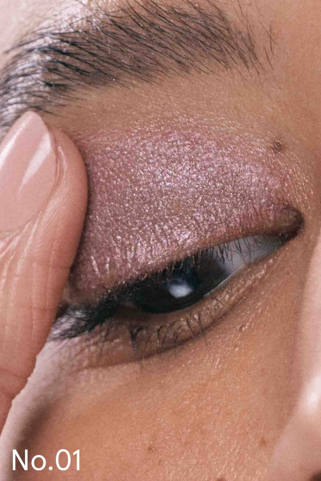 Nahaufnahme eines geschlossenen Auges mit rosa schimmerndem Lash, Lip & Cheek Trio, einer Augenbraue und einem Finger, der sanft das Augenlid berührt. Der Text No.01 erscheint in der unteren linken Ecke.