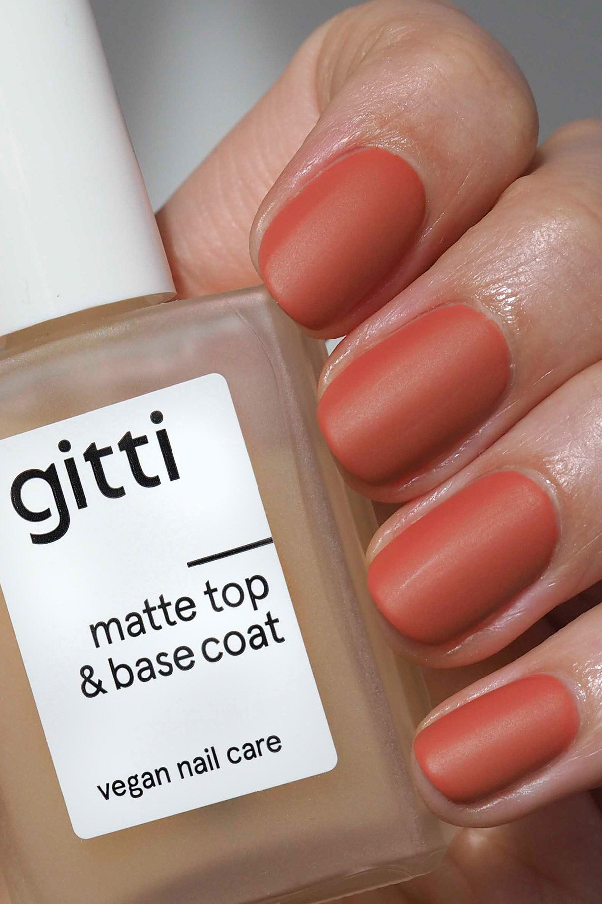 Eine Hand mit kurzen, mandelförmigen Nägeln in einem matten Terrakotta-Ton hält eine Flasche mit der Aufschrift Top & Base Coat Duo-vegane Nagelpflege für kreative und lang anhaltende Looks.