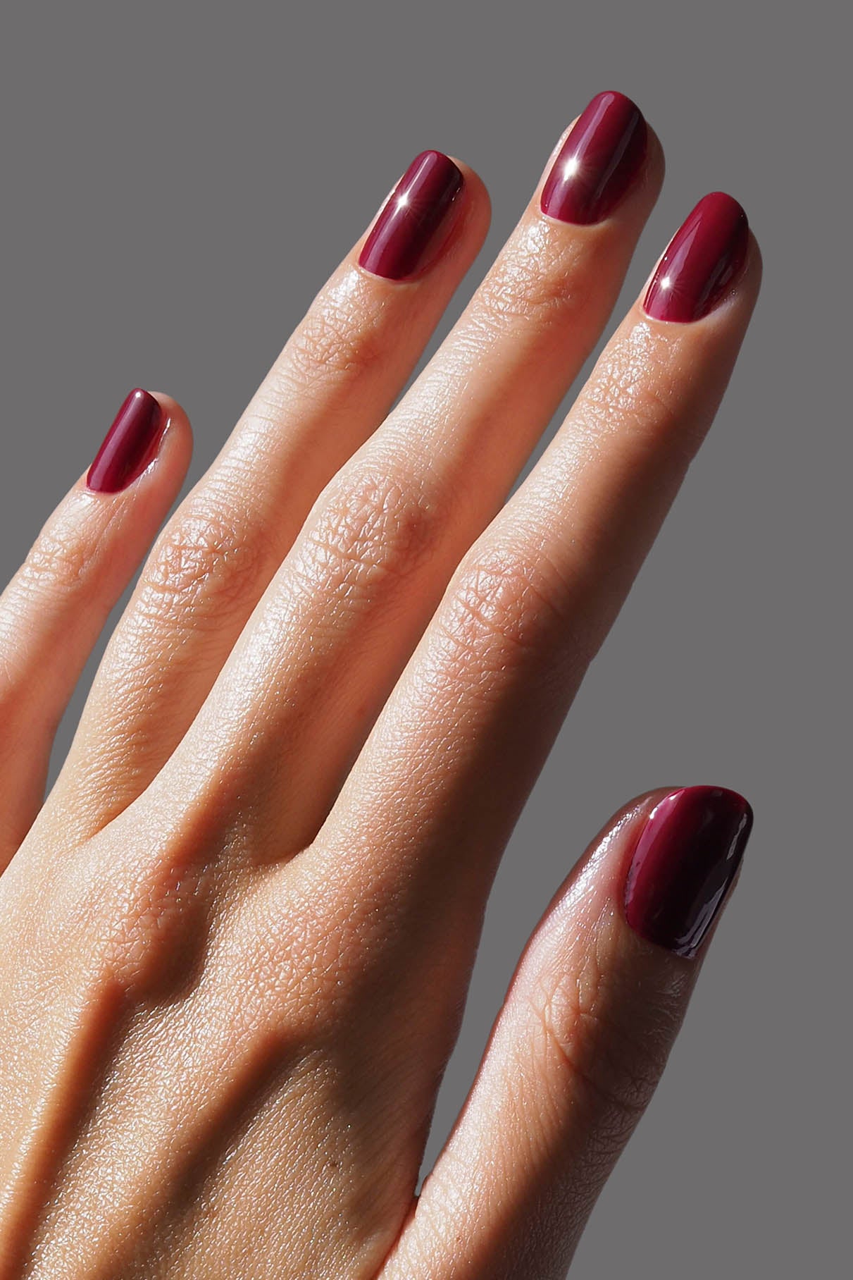 Nahaufnahme einer Hand mit kurzen, sauber manikürten Nägeln, die in Gitti Nr. 185, einem tiefen Rot mit kühlen Untertönen, vor einem schlichten grauen Hintergrund.