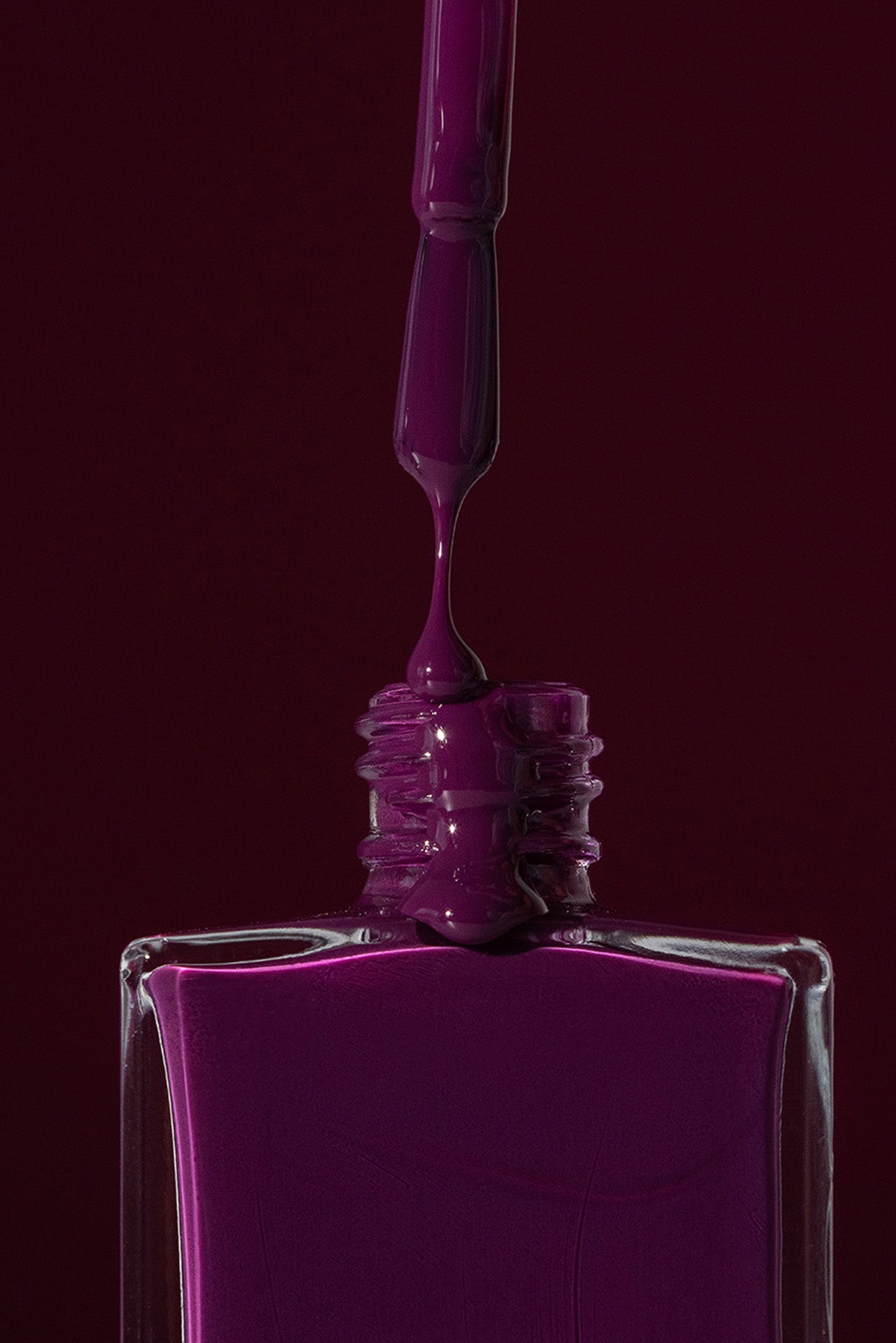 Eine Nahaufnahme eines Pinsels, der lila Gitti No. 186 Nagellack in eine viereckige Flasche tropft, vor einem dunkelvioletten und smaragdgrünen Hintergrund.