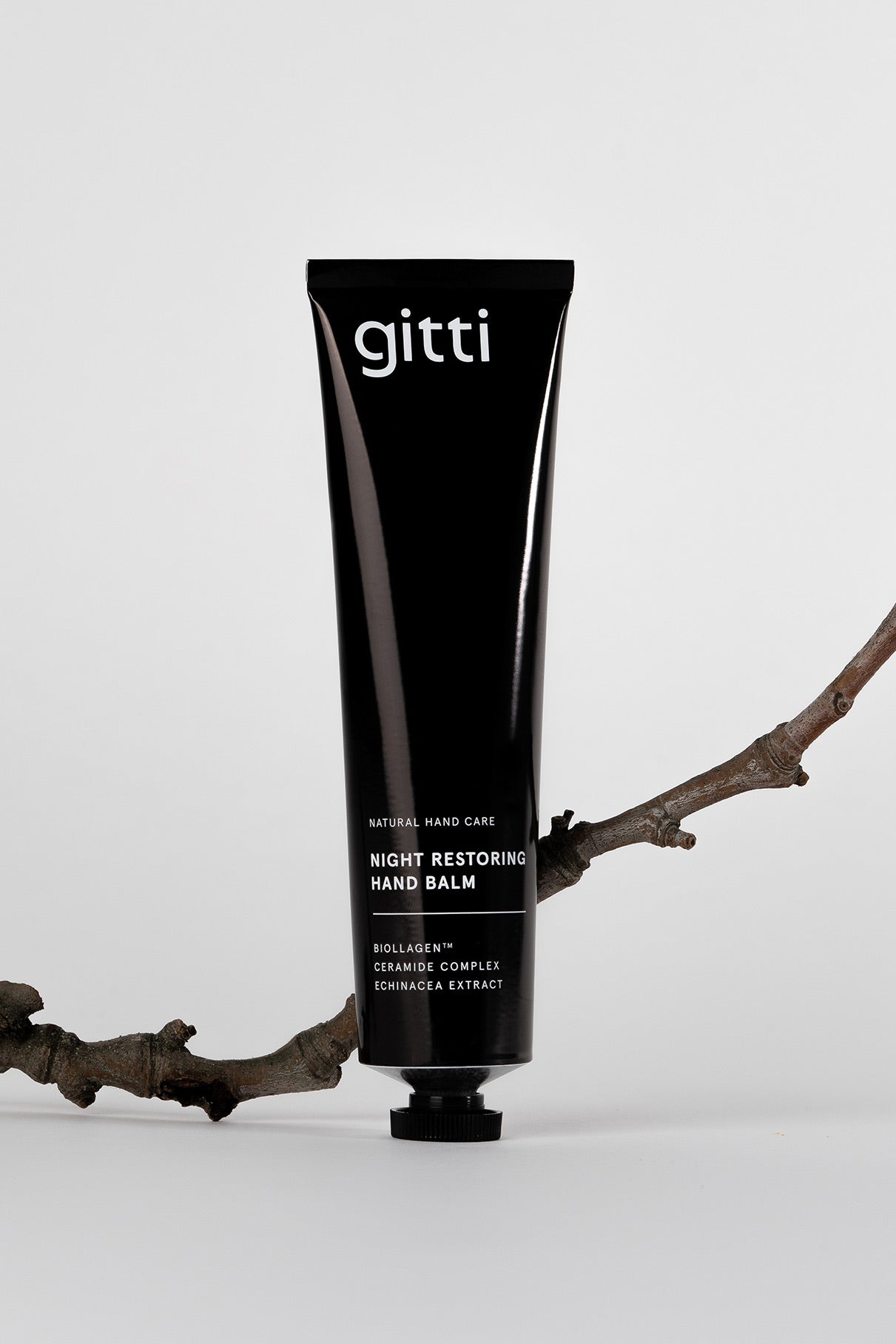 Eine schwarze Tube des Night Restoring Hand Balm steht aufrecht auf einem weißen Hintergrund, gestützt von einem trockenen Zweig - die ideale Pflege für Ihre Hände über Nacht.