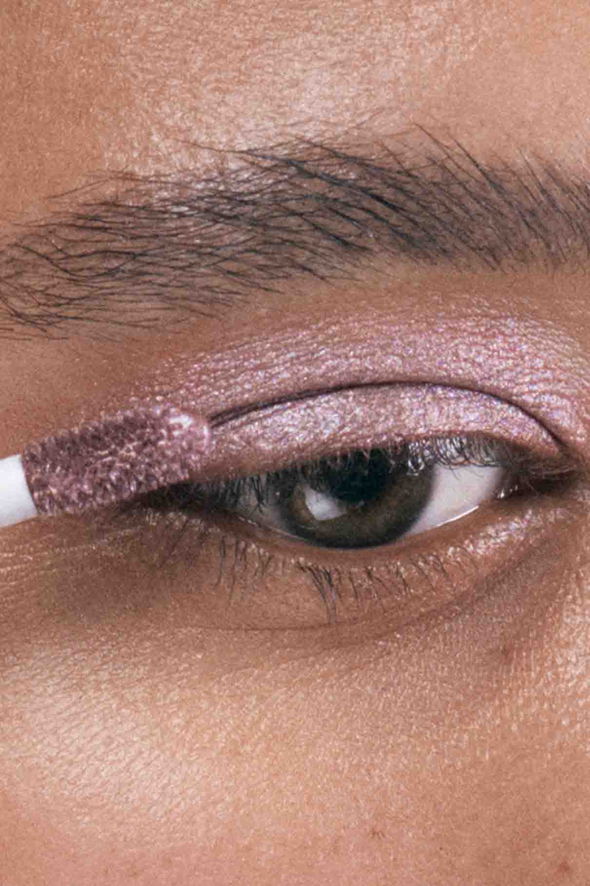 Eine Nahaufnahme eines Auges, während Visionist Nr. 01, ein schimmernder, beerenfarbener Lidschatten mit kühlem Unterton, mit einem Make-up-Applikator auf das Augenlid aufgetragen wird.