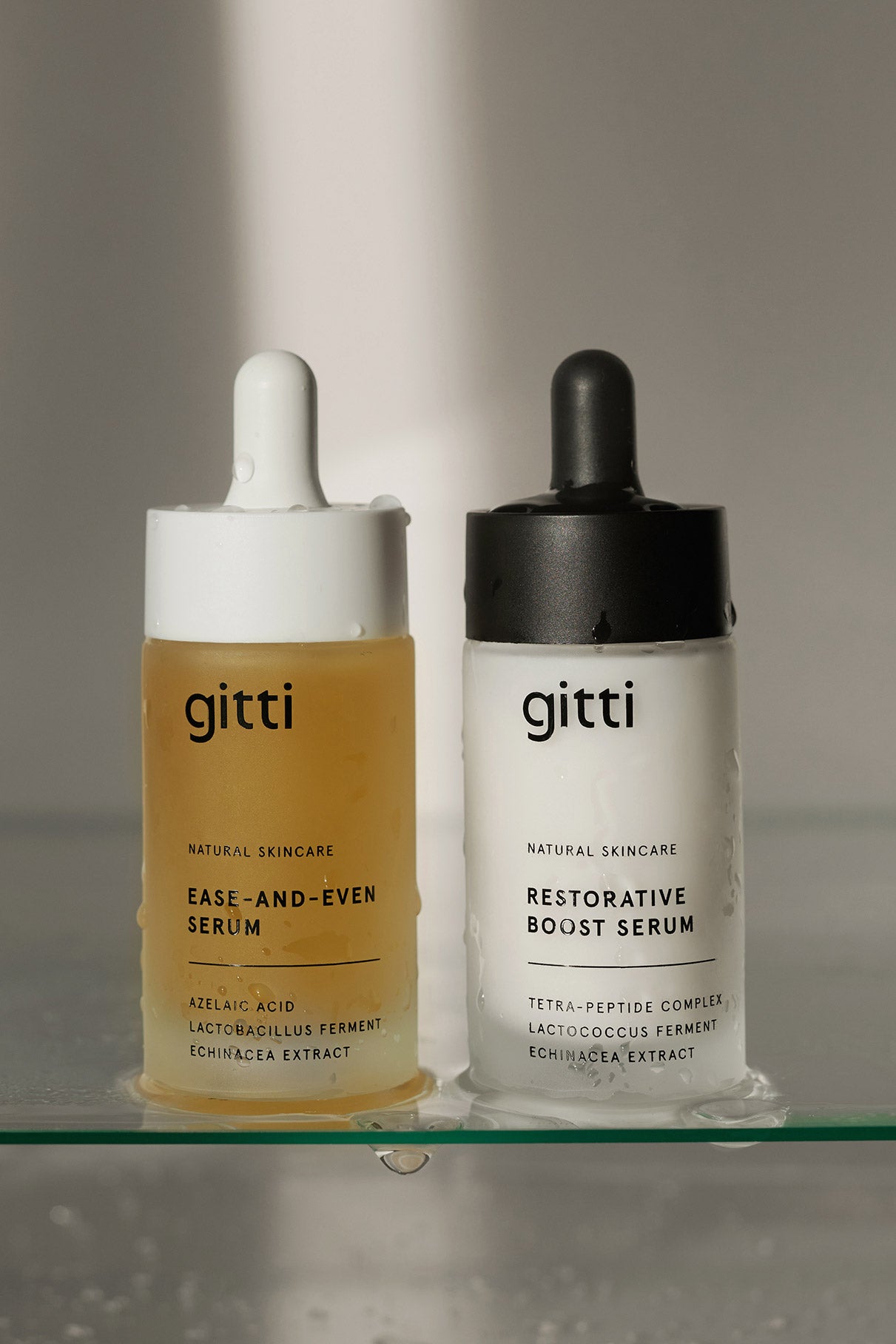 Zwei Serum-Duo-Flaschen mit der Aufschrift gitti stehen nebeneinander auf einem Glasregal - eine gelblich, eine weiß, beide mit Tropfdeckel und Produktetikett. Das perfekte Set für deine Wahl an Pflegeprodukten.