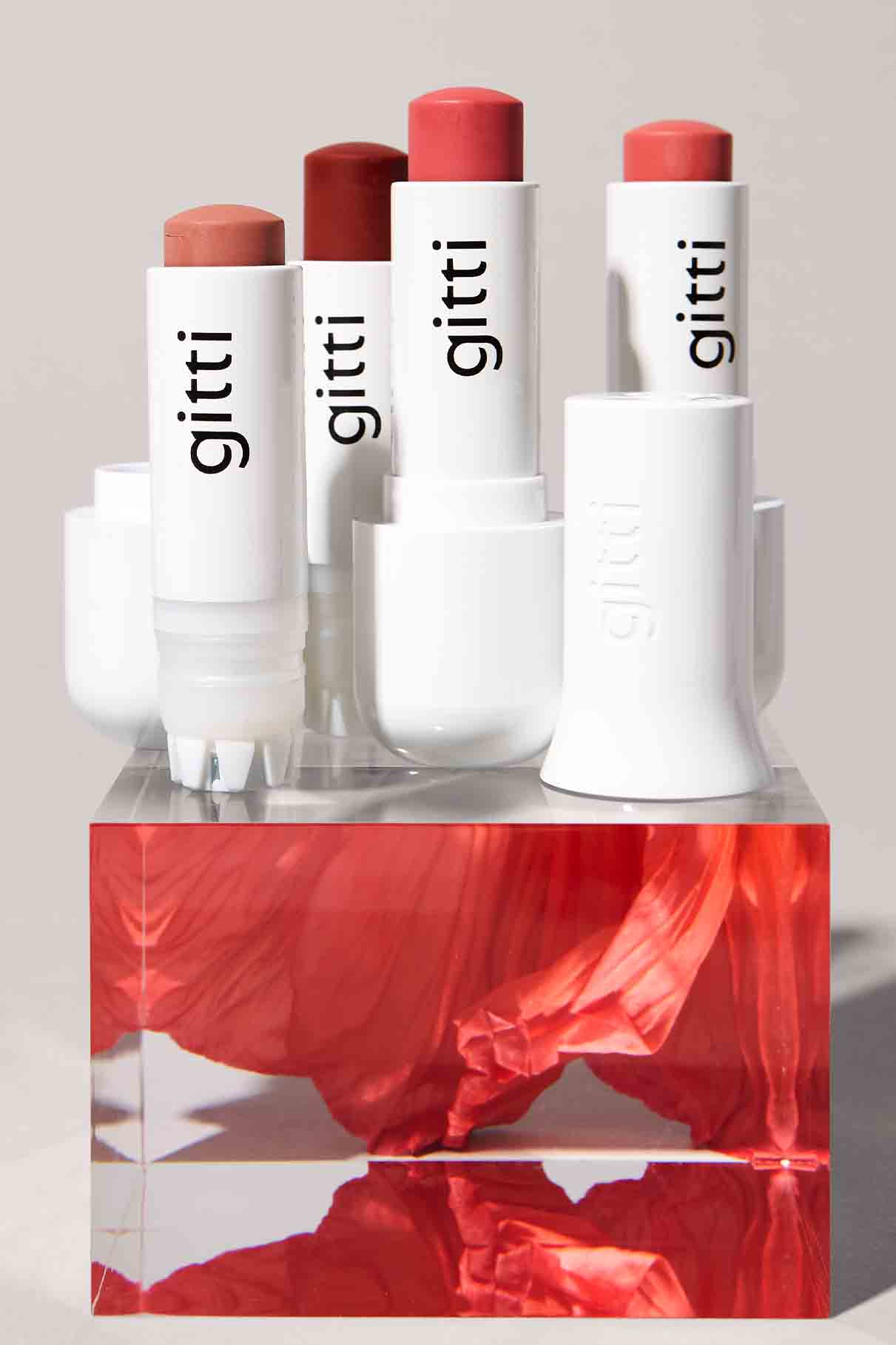 Das Set "Nails + Lips in Sync" besteht aus vier Lippenstiften der Marke gitti in weißen Tuben, die auf einem transparenten Würfel mit rotem und rosa Blumendesign präsentiert werden.