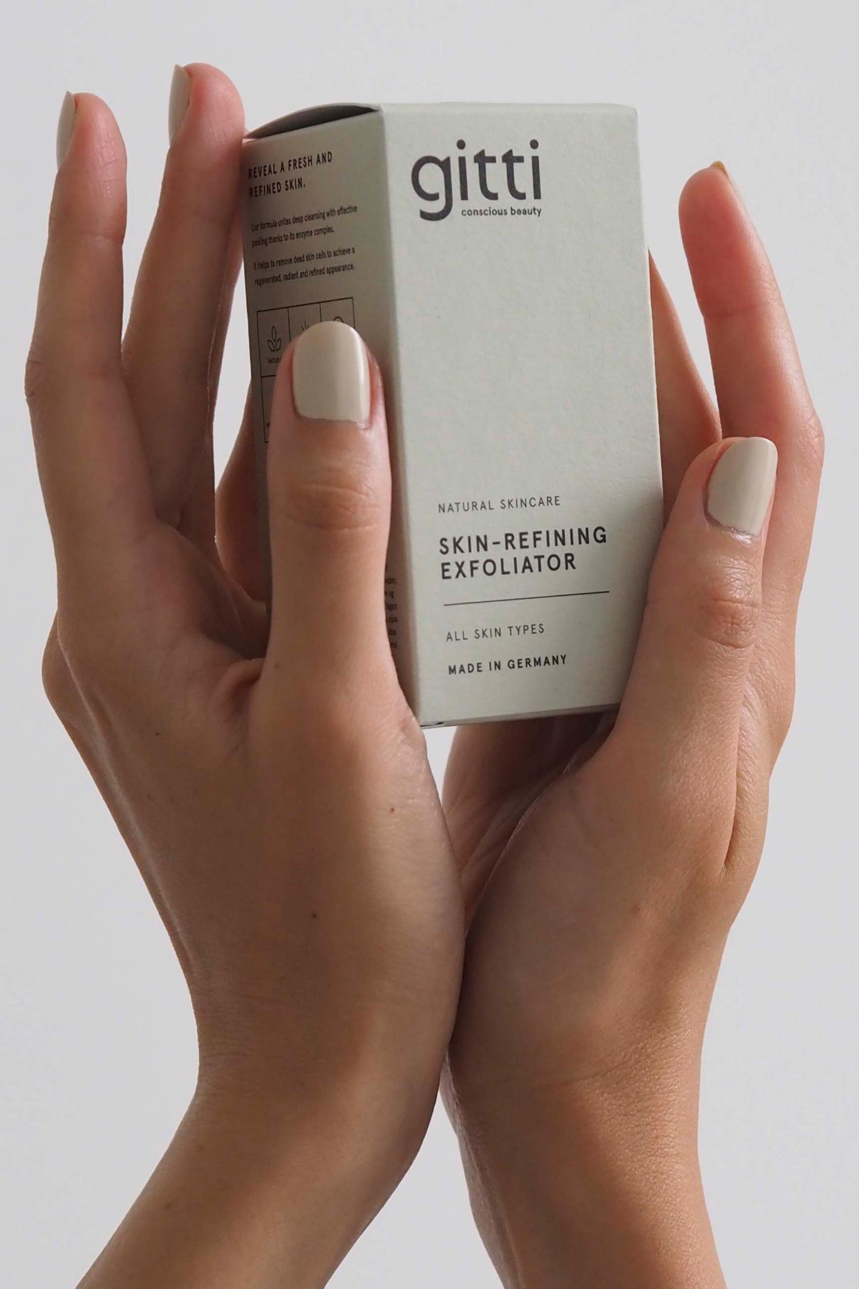 Zwei Hände mit hellem Nagellack halten eine Schachtel mit der Aufschrift Skin-Refining Exfoliator vor einem schlichten Hintergrund.
