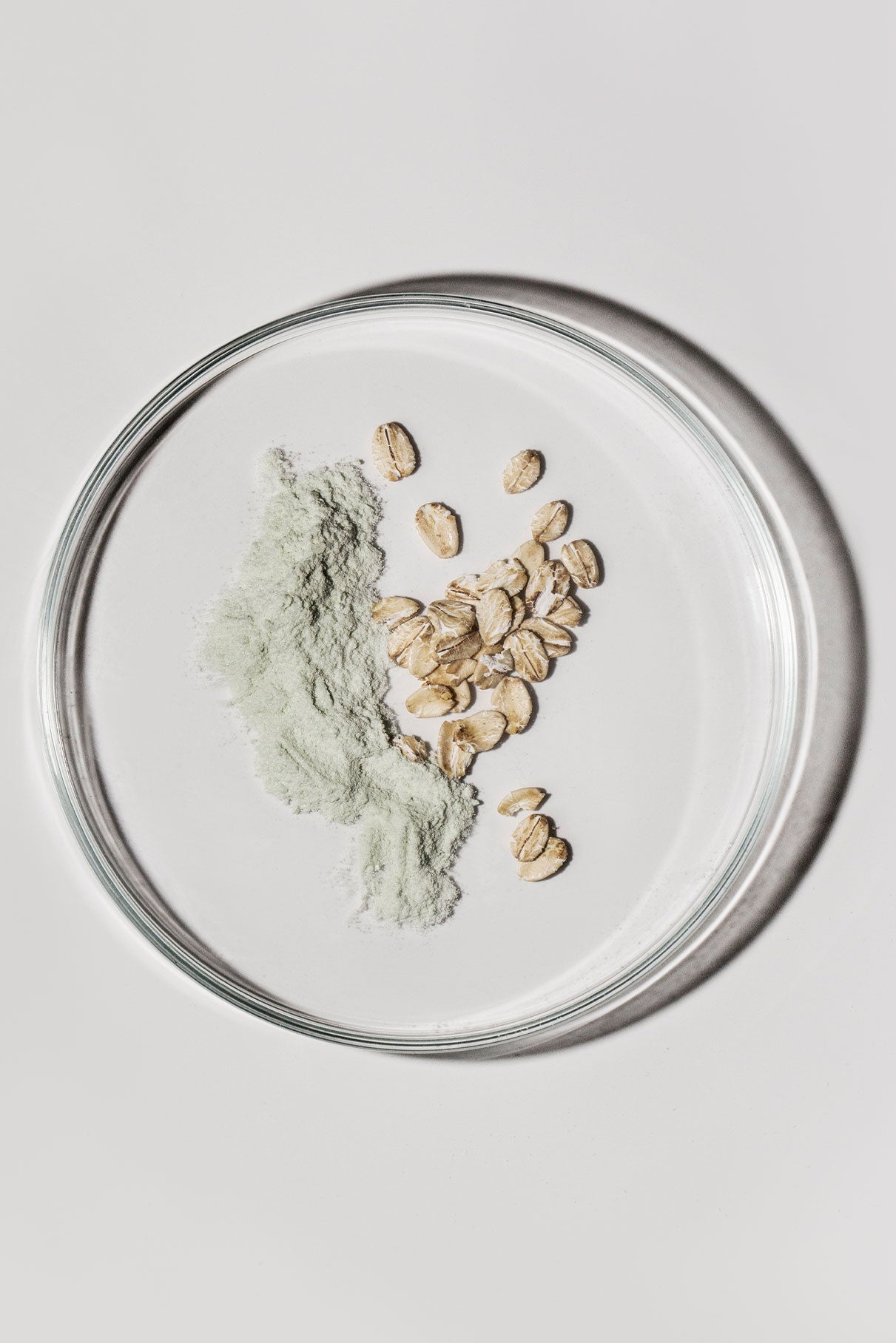 Eine gläserne Petrischale mit einem kleinen Haufen grünem Skin-Refining Exfoliator-Pulver und ganzem Hafer auf weißem Grund - ideal für die sanfte Reinigung als natürliche Peeling-Maske.