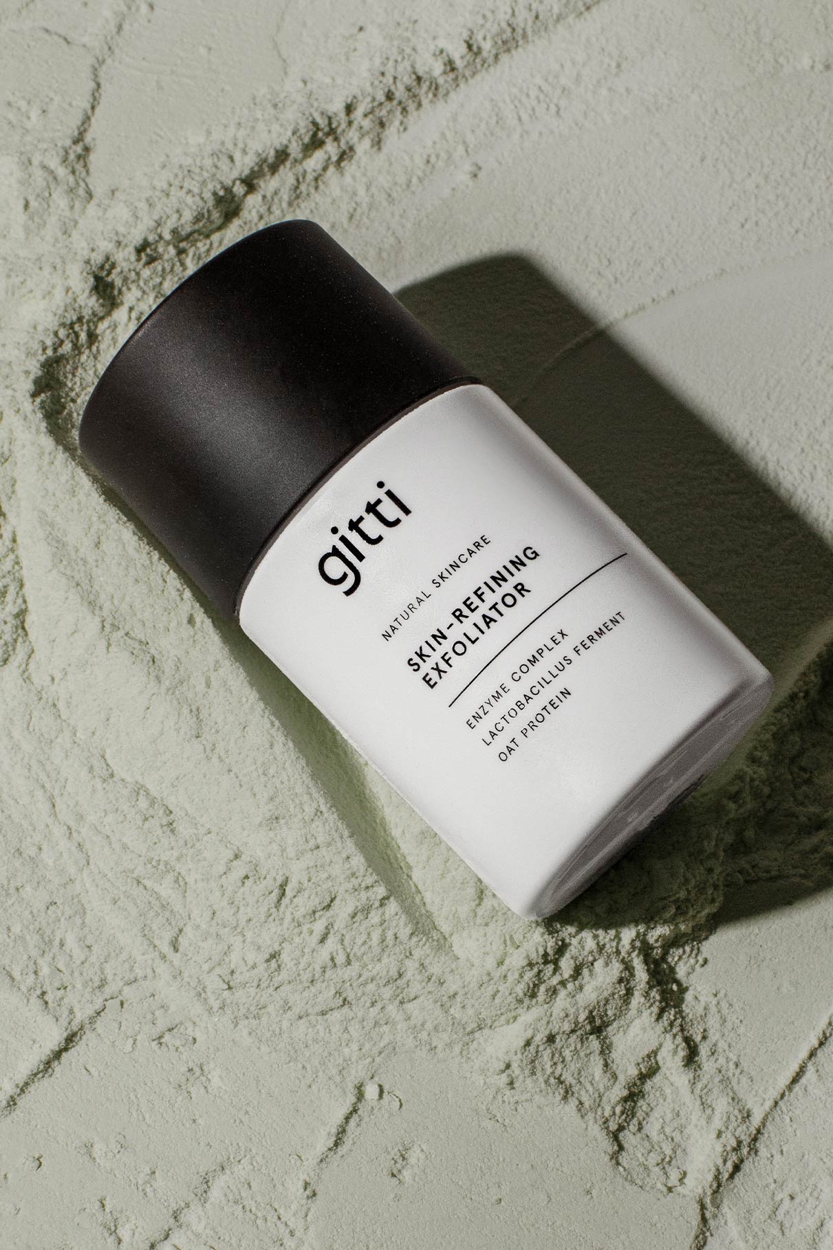 Ein weißes Fläschchen mit der Aufschrift "Skin-Refining Exfoliator" und einer schwarzen Kappe ruht auf einem strukturierten, hellgrünen Pulver - ideal für die sanfte Reinigung und die Verwendung als Enzym-Peeling oder Peeling-Maske.