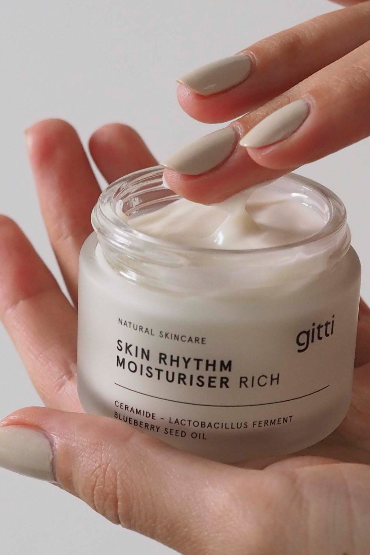 Eine Hand hält einen geöffneten Tiegel Skin Rhythm Moisturiser Rich, während die andere Hand einen Finger in die Creme taucht, die intensive Feuchtigkeit spendet und ein gesundes Strahlen hervorhebt.