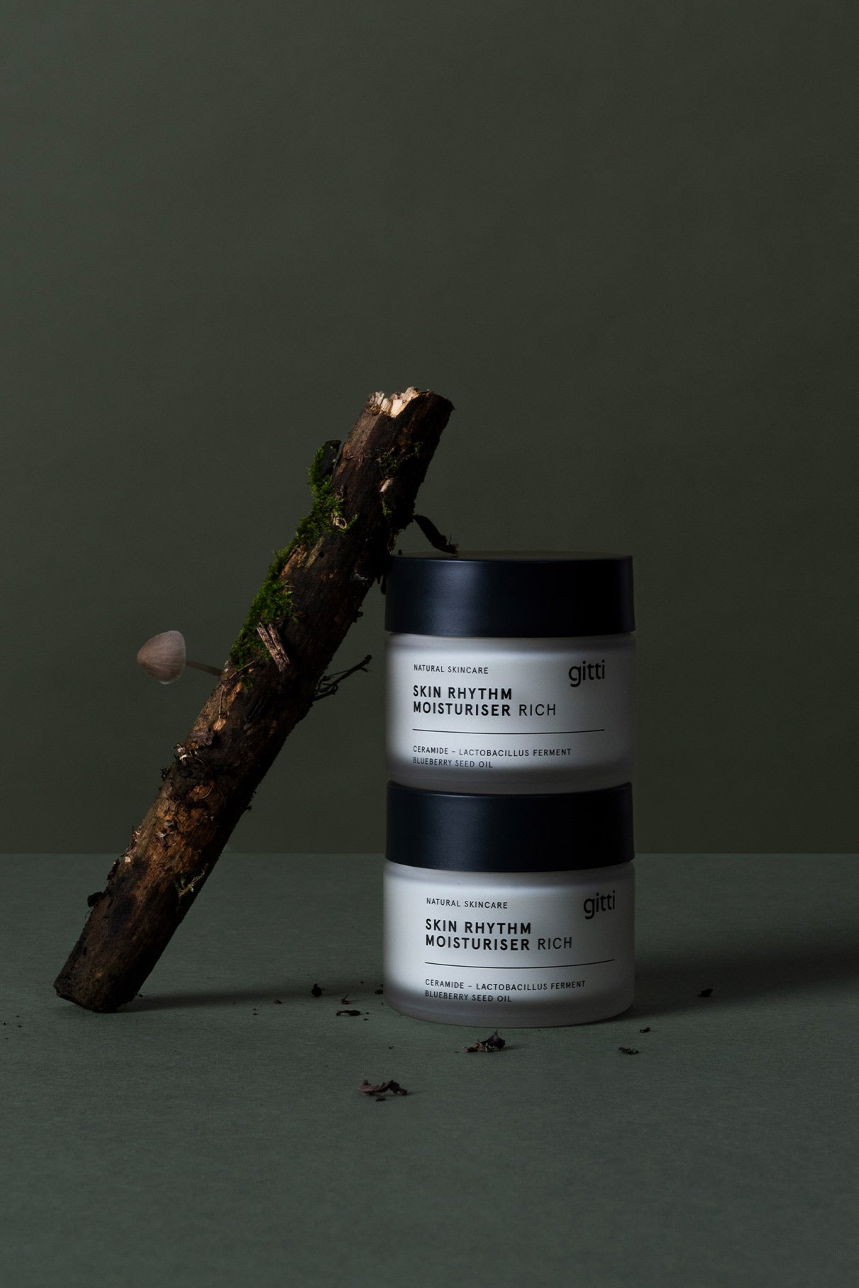 Zwei Tiegel Skin Rhythm Moisturiser Rich sind neben einem moosbewachsenen Zweig und einem Pilz gestapelt und betonen die reichhaltige Feuchtigkeit und den Barriereschutz vor einem schlichten dunklen Hintergrund.